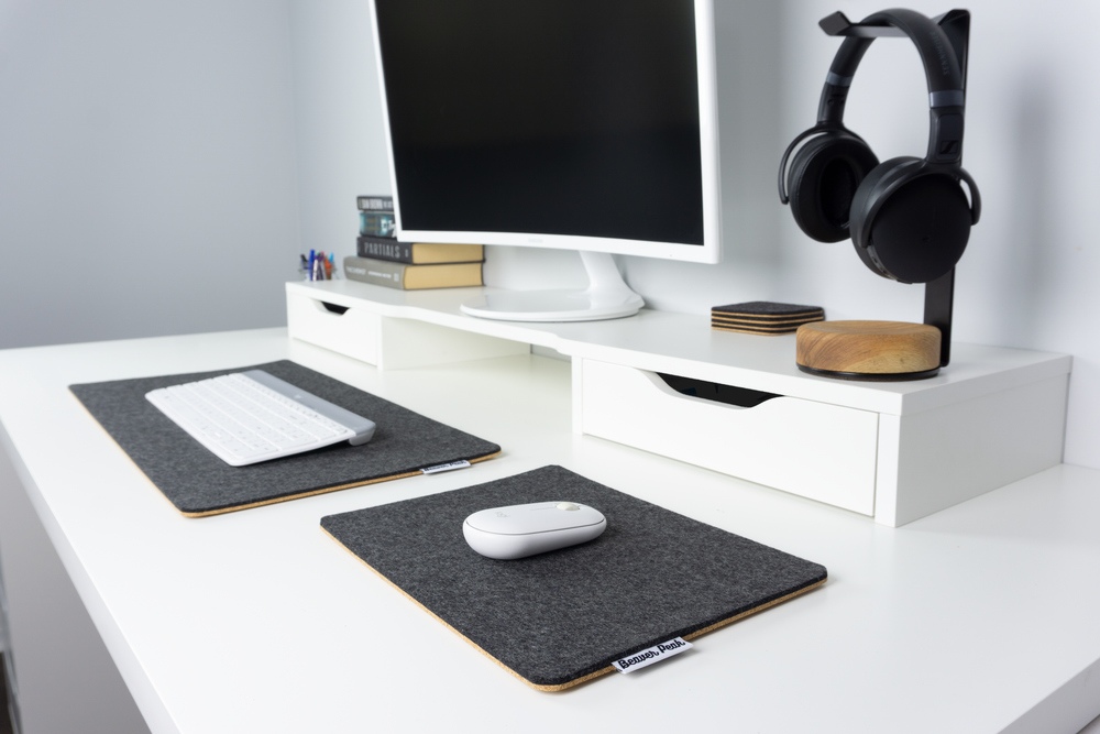 A desk setup with BeaverPeak desk products. From the left we have a black wool felt and cork desk mat, a matching black wool mouse pad and a large white desk shelf in the rear. The desk shelf holds a white monitor and our wood headphone stand plus a set of black wool coasters.